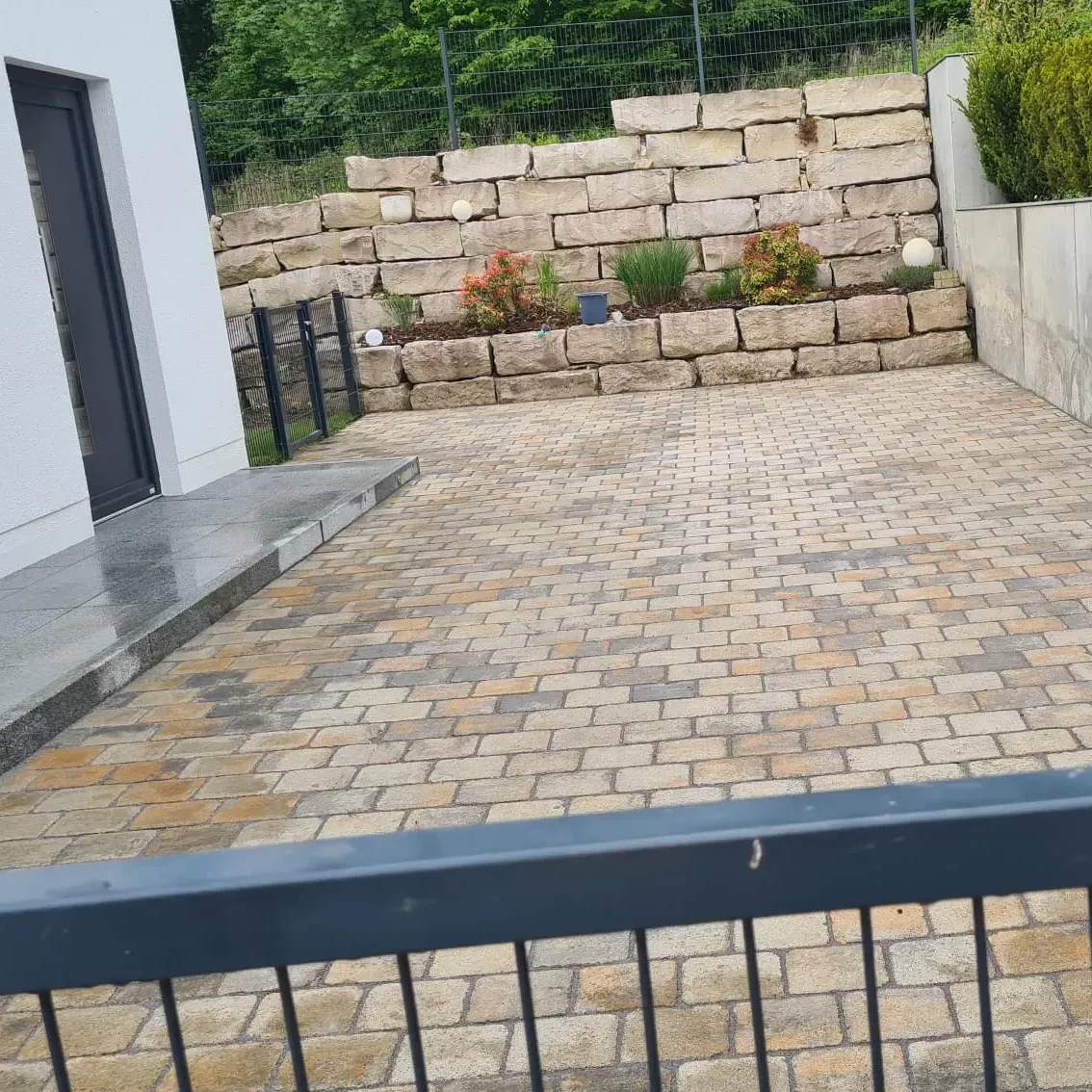 Eine mit Steinfliesen gepflasterte Auffahrt führt zu einem Schrankentor. Im Hintergrund ist eine Stützmauer aus großen Steinblöcken zu sehen, auf der kleine Sträucher und Pflanzen wachsen. Links ist teilweise ein weißes Gebäude mit einem dunklen Fensterrahmen zu sehen.