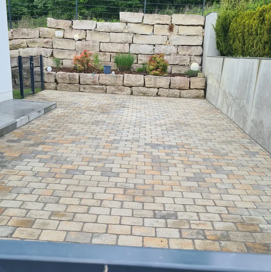 Eine mit rechteckigen Steinfliesen gepflasterte Auffahrt, die am Ende von einer niedrigen Steinmauer begrenzt wird. Entlang der Mauer sind kleine Pflanzen und Sträucher zu sehen. Auf der rechten Seite befindet sich eine graue Betonmauer mit Grünpflanzen darüber.
