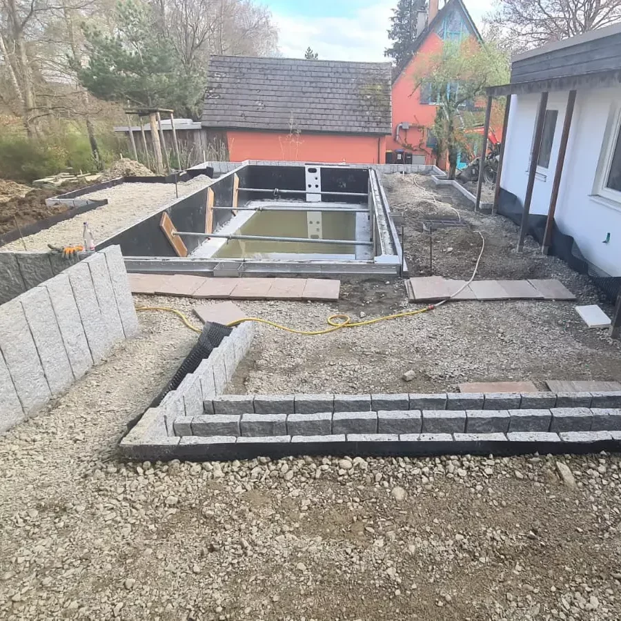 Ein im Bau befindlicher Hinterhof mit einem teilweise gebauten, mit Wasser gefüllten Pool. Um den Pool herum sind Steinplatten und Kieswege. Im Hintergrund sind Bäume und ein Haus mit einer orangefarbenen Wand. Auf der rechten Seite ist ein weißes Gebäude.