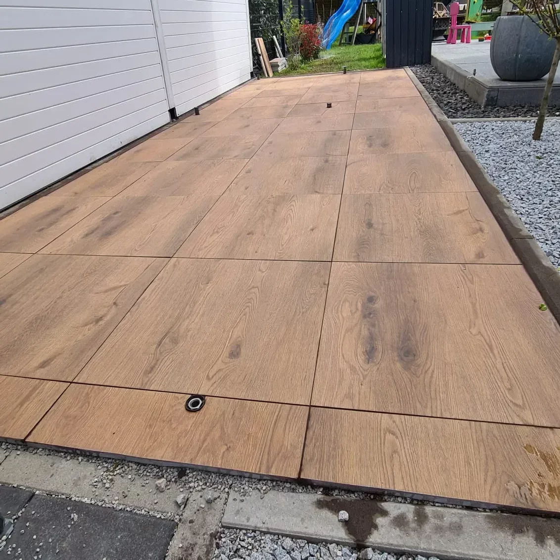 Eine neu angelegte Terrasse mit großen Fliesen in Holzoptik erstreckt sich an der Seite eines weißen Gebäudes. Bei den grünen Pflanzen im Hintergrund liegen verstreut Kinderspielzeuge herum. Der Rand der Terrasse ist mit Kies gesäumt.