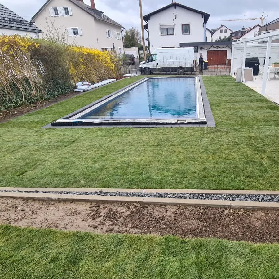Ein moderner rechteckiger Außenpool ist von frisch verlegtem grünem Gras in einem Vorstadtgarten umgeben. Im Hintergrund sind zwei Häuser und ein geparkter Lieferwagen unter einem bewölkten Himmel zu sehen. Eine Reihe gelber Sträucher säumt den Rand des Gartens.